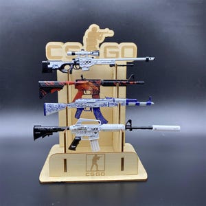 May include: A wooden display stand featuring the CS:GO logo and silhouette, holding four miniature toy rifle models. The rifles include a white sniper rifle, a red and black patterned rifle, a blue and white floral rifle, and a white rifle with a silencer.
