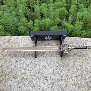 Pu&ograve; includere: Una spada decorativa con una lama in bronzo e un'elsa ornata, esposta su un supporto in legno nero. L'elsa presenta dettagli intricati e un'impugnatura marrone scuro. La spada &egrave; lunga circa 71 cm.