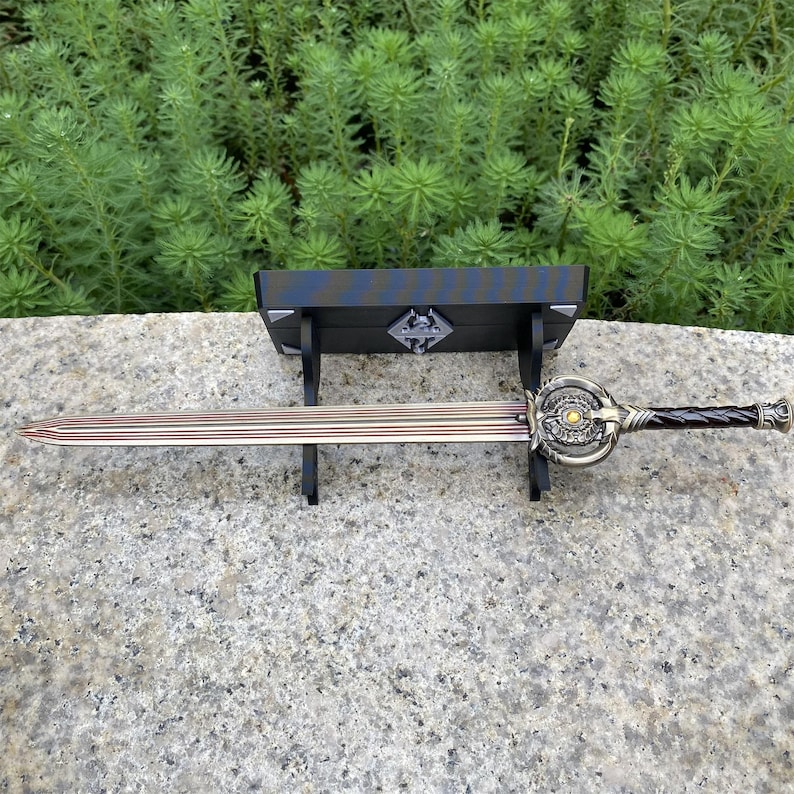 Pu&ograve; includere: Una spada decorativa con una lama in bronzo e un'elsa ornata, esposta su un supporto in legno nero. L'elsa presenta dettagli intricati e un'impugnatura marrone scuro. La spada &egrave; lunga circa 71 cm.