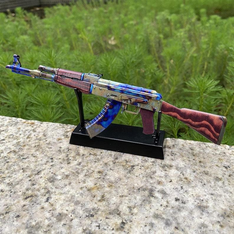 May include: A detailed model of an AK-47 assault rifle, featuring a vibrant color scheme of blues, greens, and reds. The rifle is mounted on a black stand, showcasing its intricate design and craftsmanship. The stock and handguard have a wood-grain appearance.