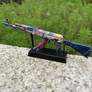 May include: A decorative AK-47 rifle model, featuring a detailed design with blue, red, and gold accents. The model is displayed on a black stand, showcasing intricate patterns and embellishments. The rifle is approximately 30 inches long.
