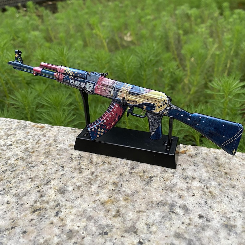 May include: A decorative AK-47 rifle model, featuring a detailed design with blue, red, and gold accents. The model is displayed on a black stand, showcasing intricate patterns and embellishments. The rifle is approximately 30 inches long.
