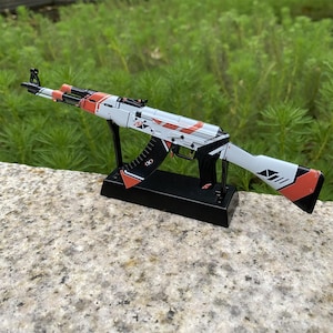 May include: A detailed model of an AK-47 assault rifle, featuring a white, black, and orange color scheme. The model is displayed on a black stand, showcasing its design. The rifle's barrel and magazine are black, with orange accents.