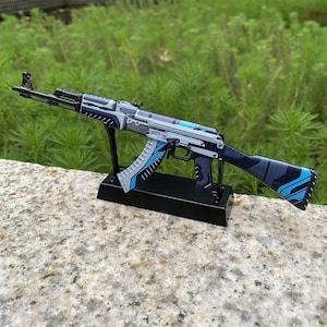 May include: A detailed model of an AK-47 assault rifle, featuring a black, gray, and blue color scheme. The rifle is mounted on a black stand, showcasing its design and intricate details. The background is a blurred green.