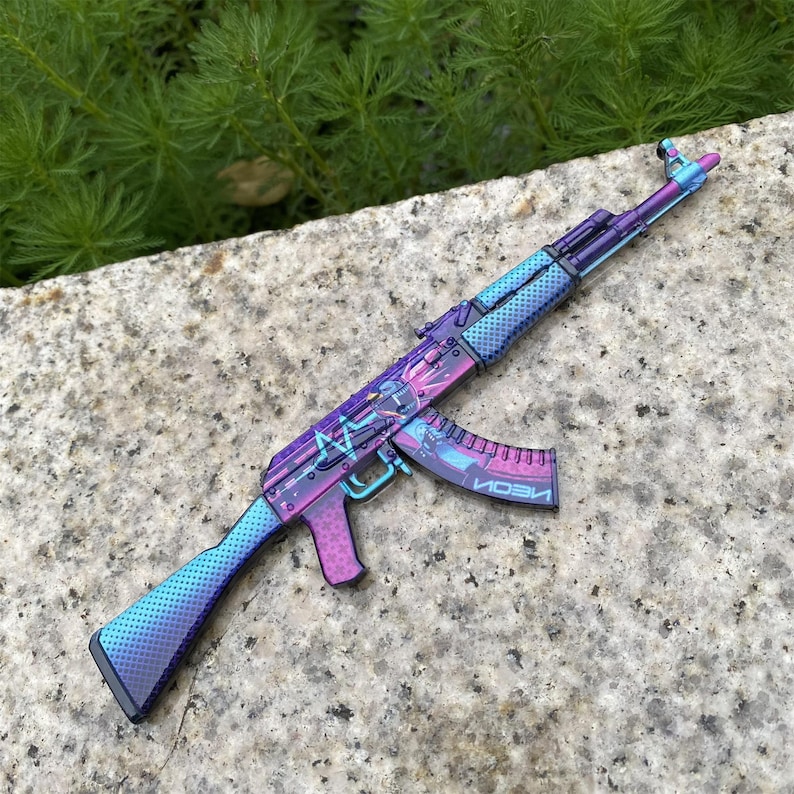 May include: A colorful AK-47 rifle with a gradient design in shades of purple, blue, and pink. The magazine reads "NEON" in a futuristic font. The gun is resting on a stone surface.