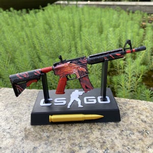 Op de afbeelding: Een decoratief speelgoedgeweer met een rood en zwart ontwerp, gepresenteerd op een zwarte standaard. De standaard toont de tekst "CS GO" en een goudkleurige kogel. Het geweer heeft een rode grip en accenten.
