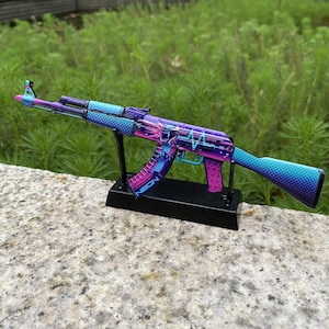 May include: A colorful AK-47 replica with a gradient design of blue, purple, and pink. The gun features a black stand and the word "NEON" is visible on the magazine. The design includes a heartbeat graphic and other neon elements.