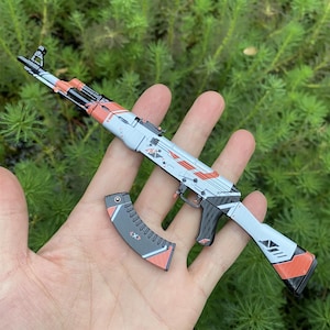 May include: A miniature AK-47 style toy gun with a detached magazine. The toy gun is primarily light gray with orange and black accents. The magazine is also light gray with orange and black details. The toy is held in a hand against a green background.