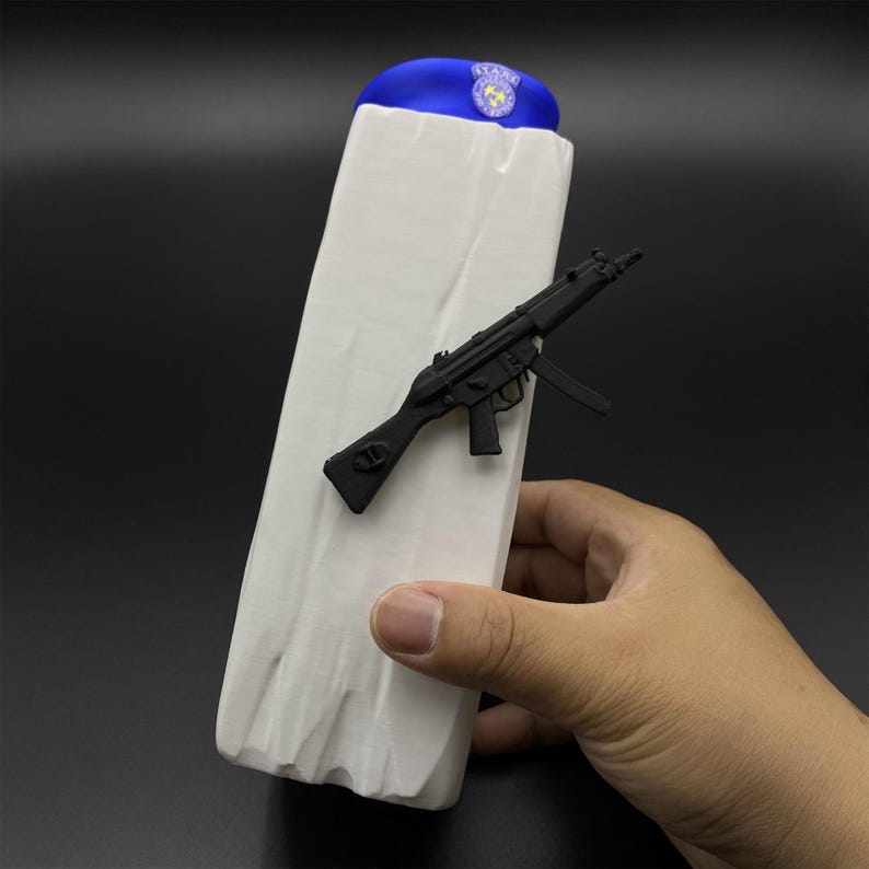 May include: A white rectangular object with a blue beret on top and a black toy gun attached. The beret has a badge. The object is held by a hand.
