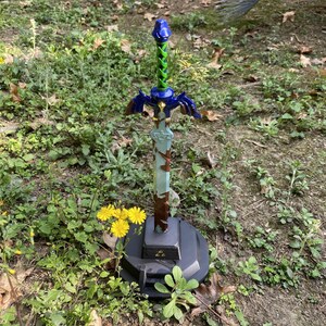 May include: A decorative sword replica with a blue and green handle, blue crossguard, and a light blue blade. The sword is mounted on a black base with a gold triangle emblem. The sword is displayed outdoors among green plants and yellow flowers.