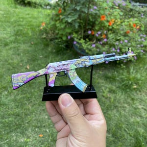 Könnte beinhalten: Eine farbenfrohe AK-47-Replik mit einem lebendigen, gemusterten Design in Lila, Blau und Gelb. Die Spielzeugpistole ist auf einem schwarzen Ständer montiert, der ihre komplizierten Details hervorhebt. Der Hintergrund zeigt grünes Gras und Blätter.