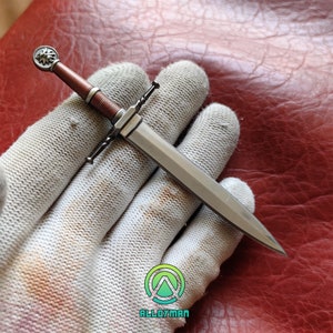 May include: A silver and brown miniature sword with a detailed hilt featuring a circular emblem. The handle is wrapped in brown material, and the blade is sharp and pointed. The sword is held against a white glove.