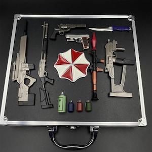 May include: A collection of miniature toy weapons and accessories, including guns, a knife, grenades, and an umbrella-shaped emblem with red and white segments. The items are arranged on a black surface within a silver-edged case.