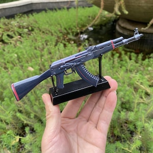 Könnte beinhalten: Ein detailliertes Miniaturmodell eines AK-47-Gewehrs, überwiegend schwarz mit roten Akzenten an Magazin, Schaft und Handschutz. Das Modell ist auf einem schwarzen Ständer montiert und zeigt sein kompliziertes Design und seine realistischen Merkmale.