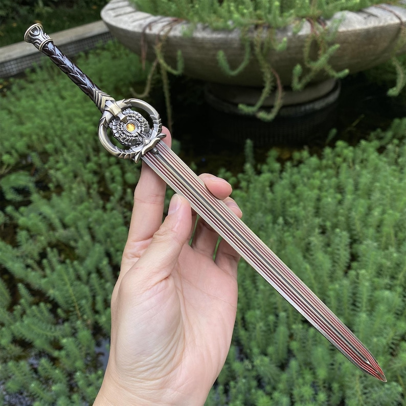 Pu&ograve; includere: Un pugnale ornato con una lama e un'impugnatura color bronzo. L'impugnatura presenta un design dettagliato con un elemento circolare centrale e un'impugnatura intrecciata marrone scuro. La lama ha linee parallele e una punta rossa.