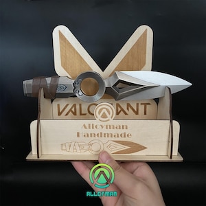May include: A handmade, metallic dagger with a brown leather-wrapped handle, displayed on a wooden stand. The stand features the word "VALORANT" and "Alloyman Handmade" engraved on it. The dagger has a silver blade and a circular design element.