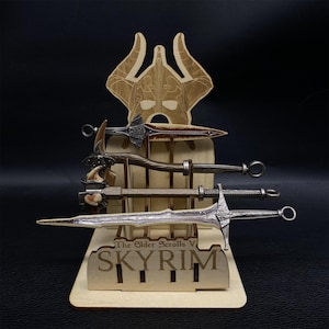 May include: A wooden display stand featuring four miniature weapons, including swords and a hammer. The stand has a decorative top with a helmet design and the text "The Elder Scrolls V SKYRIM" at the base. The weapons are various shades of brown and silver.