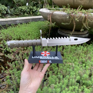 Op de afbeelding: Een zilverkleurig bajonetmes met een gekartelde rand en een getextureerd handvat. Het mes staat op een zwarte standaard met het Umbrella Corporation logo en de tekst "Resident Evil".