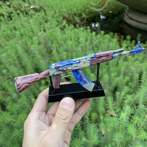 May include: A detailed model of an AK-47 assault rifle. The model features a colorful, iridescent finish with blue, purple, and gold hues. The wooden stock and handguard have a reddish-brown wood grain pattern. The model is mounted on a black stand.