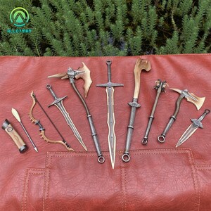 Puede incluir: Una colecci&oacute;n de armas de fantas&iacute;a en miniatura, que incluye espadas, hachas, un arco y una flecha, y una maza. Las armas tienen detalles met&aacute;licos y de madera, dispuestas sobre una superficie roja similar al cuero. El fondo presenta follaje verde.