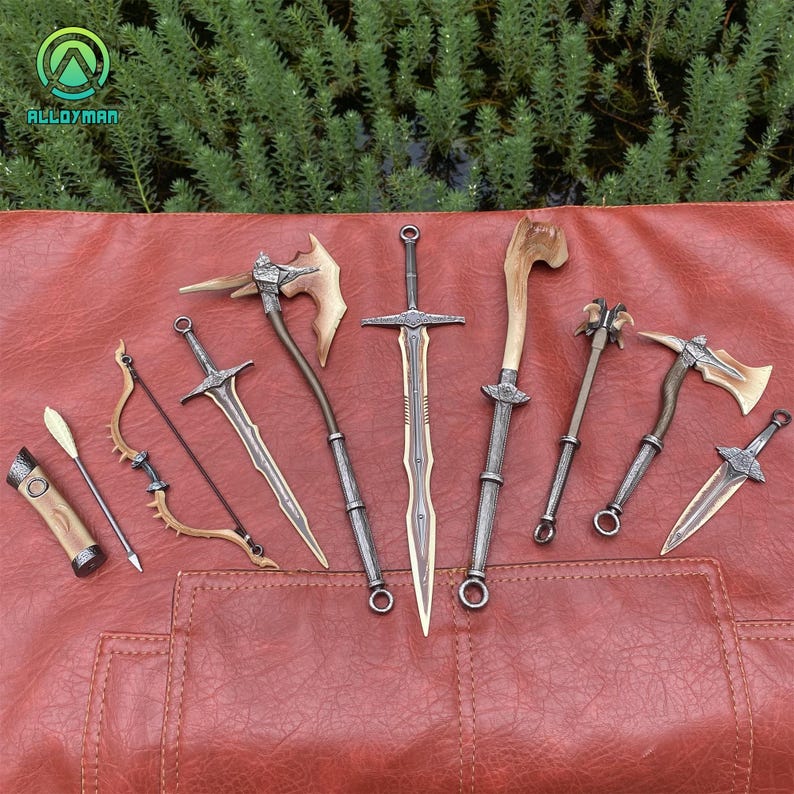 Puede incluir: Una colecci&oacute;n de armas de fantas&iacute;a en miniatura, que incluye espadas, hachas, un arco y una flecha, y una maza. Las armas tienen detalles met&aacute;licos y de madera, dispuestas sobre una superficie roja similar al cuero. El fondo presenta follaje verde.