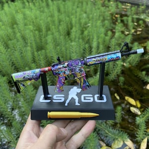 May include: A colorful toy rifle with a graffiti-style design, displayed on a black stand with the text "CS GO" and a gold bullet. The rifle features a variety of colors, including purple, green, and red.