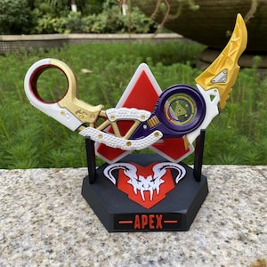 May include: A gold, white, and purple karambit knife replica with a red and white triangle and a black base. The base has the word "APEX" in white text. The knife has a circular handle and a curved blade.