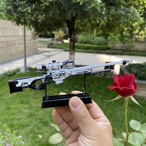 Peut inclure: Un fusil de sniper miniature, aux couleurs noir et blanc, avec des motifs détaillés et une lunette. Il est présenté sur un support noir, devant un fond vert flou, avec une rose rouge au premier plan. Le design du fusil est complexe.