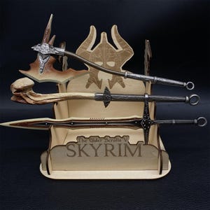 May include: A wooden display stand featuring three fantasy weapons: an axe, a sword, and a spear. The stand is engraved with the text "The Elder Scrolls V SKYRIM". The weapons have metallic and brown accents.