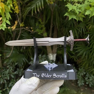 May include: A silver-colored sword with a red handle and ornate detailing, displayed on a black stand. The stand features the text "The Elder Scrolls" and a logo. The sword is a replica of a fantasy weapon.