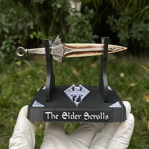 May include: A decorative dagger with a cream-colored blade and brown accents, displayed on a black stand. The stand features the text "The Elder Scrolls" and a game logo. The dagger has a silver handle and a decorative silver guard.