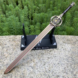 Pu&ograve; includere: Una spada decorativa con una lama argentata con dettagli rossi e dorati. L'elsa presenta un design dettagliato con un centro giallo. La spada poggia su un supporto nero con la scritta "The Elder Scrolls".