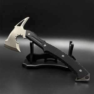 Pode incluir: Um machado tático tomahawk prateado e preto com um cabo texturizado. A cabeça do machado apresenta uma lâmina, um pico e um recorte circular. O cabo é preto com rebites prateados. O machado é exibido em um suporte preto.