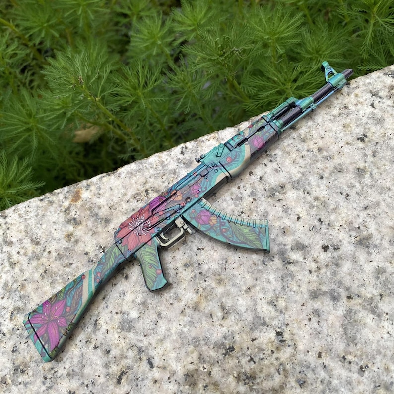 May include: A colorful AK-47 rifle with a floral design in shades of teal, purple, and pink. The firearm is resting on a gray stone surface, with green foliage in the background. The magazine is also decorated with the same floral pattern.