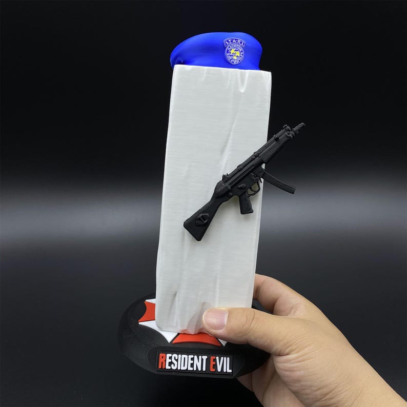 May include: A blue beret with a badge, a black toy gun, and a white rectangular object are displayed on a black base with the text "RESIDENT EVIL". The base has a red and white emblem.