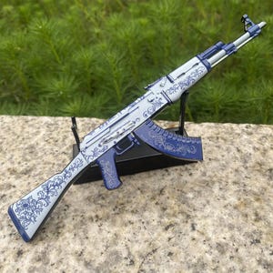 May include: A decorative AK-47 rifle model in shades of white and blue, featuring intricate floral scrollwork. The model is displayed on a black stand against a natural background. The rifle's design includes a blue magazine and grip.