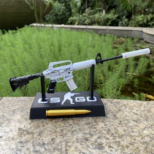 May include: A white and black toy rifle with a silencer, displayed on a black stand with the text "CS GO" and a golden bullet. The rifle has intricate detailing and is supported by two black stands.