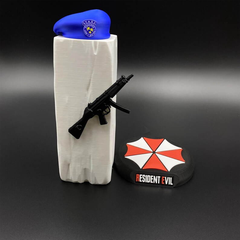 May include: A blue beret with a gold emblem, a black toy gun, and a black and red patch with the words "Resident Evil". The items are displayed on a white rectangular stand.