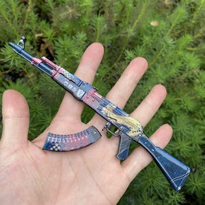 May include: A decorative AK-47 rifle with a detached magazine. The rifle features a blue, red, and gold design with a patterned finish. The magazine has a matching design. The background is a blurred green.