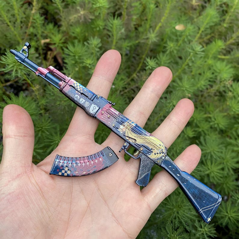 May include: A decorative AK-47 rifle with a detached magazine. The rifle features a blue, red, and gold design with a patterned finish. The magazine has a matching design. The background is a blurred green.