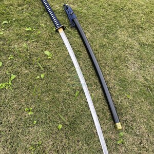 Może przedstawiać: Japoński miecz katana ze srebrnym ostrzem i czarną pochwą. Rękojeść jest owinięta niebieską tkaniną z białymi akcentami. Miecz leży na zielonej trawie.