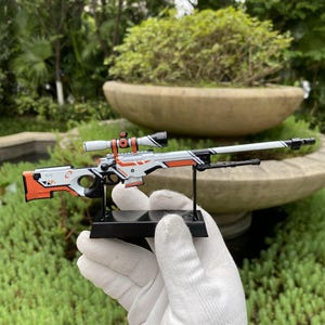 Op de afbeelding: Een miniatuur wit, oranje en zwart sluipschuttergeweer met een vizier. Het geweer is gemonteerd op een zwarte standaard. De achtergrond toont groen gebladerte en een stenen plantenbak.