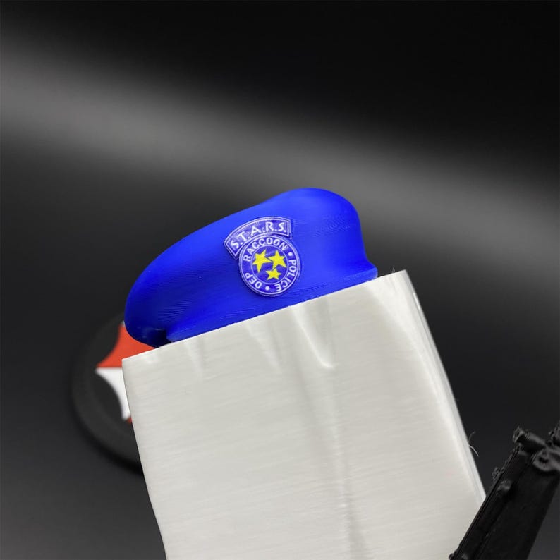 May include: A blue beret with a circular patch featuring the text "S.T.A.R.S. Raccoon Police Dept" and two yellow stars. The beret is partially obscured by a white object, with a black background.