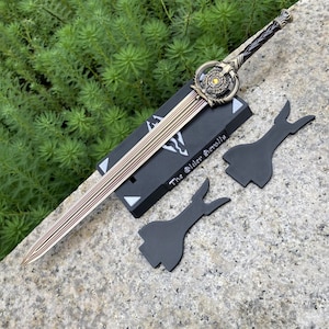 Pu&ograve; includere: Una spada decorativa con lama e impugnatura dorate, appoggiata su un supporto nero. La spada presenta un design dettagliato con una guardia circolare e un'impugnatura intrecciata. Il supporto riporta la scritta "The Elder Scrolls" e ci sono due elementi decorativi neri.