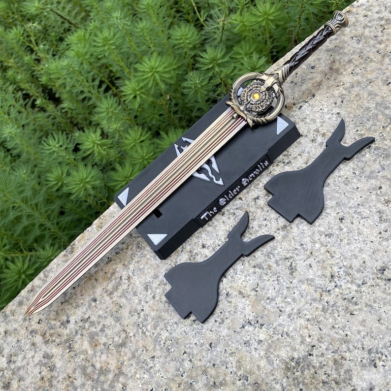 Pu&ograve; includere: Una spada decorativa con lama e impugnatura dorate, appoggiata su un supporto nero. La spada presenta un design dettagliato con una guardia circolare e un'impugnatura intrecciata. Il supporto riporta la scritta "The Elder Scrolls" e ci sono due elementi decorativi neri.