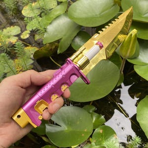May include: A decorative knife with a gold blade and a pink and gold handle. The blade has a serrated edge. The handle has a series of gold accents and a white cylindrical component. The knife is held over water with lily pads.