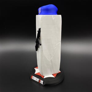 May include: A white rectangular figurine with a black gun attached to the side. A blue beret sits on top. The base is black with a red and white design and the words "RESIDENT EVIL" in white.