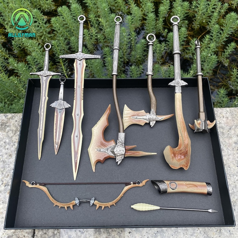 Puede incluir: Una colecci&oacute;n de armas de fantas&iacute;a en miniatura, que incluye espadas, hachas, un arco y flechas y una maza, exhibidas en una bandeja negra. Las armas presentan tonos plateados y marrones, con detalles intrincados. El logotipo "ALLOYMAN" es visible.