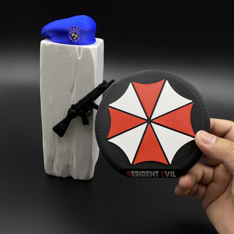 May include: A blue beret with a badge, a black toy gun, and a round patch with a red and white umbrella design and the text "RESIDENT EVIL". The items are displayed on a white stand.