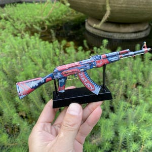 Könnte beinhalten: Ein Miniaturmodell eines AK-47-Gewehrs mit einem rot, blau und weiß gehaltenen Design. Das Modell ist auf einem schwarzen Ständer montiert und trägt das Wort "VOLATJOV" auf dem Magazin. Der Hintergrund ist eine verschwommene Ansicht von grünem Laub.
